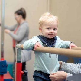 A toddler smiles at the camera whilst on parallel bars